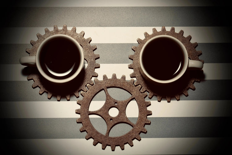Two coffee cups on metallic gear-shaped coasters with a third gear on striped surface